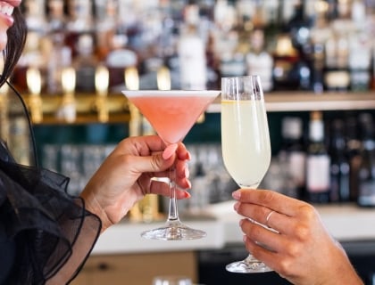 A woman raises a cocktail glass in a toast while sitting at a bar, smiling and enjoying the moment.