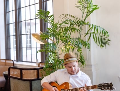 A man strumming a guitar, immersed in music, with a focused expression and a casual setting around him.