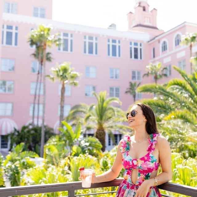 A woman in a floral jumpsuit drinking a cocktail on a balcony