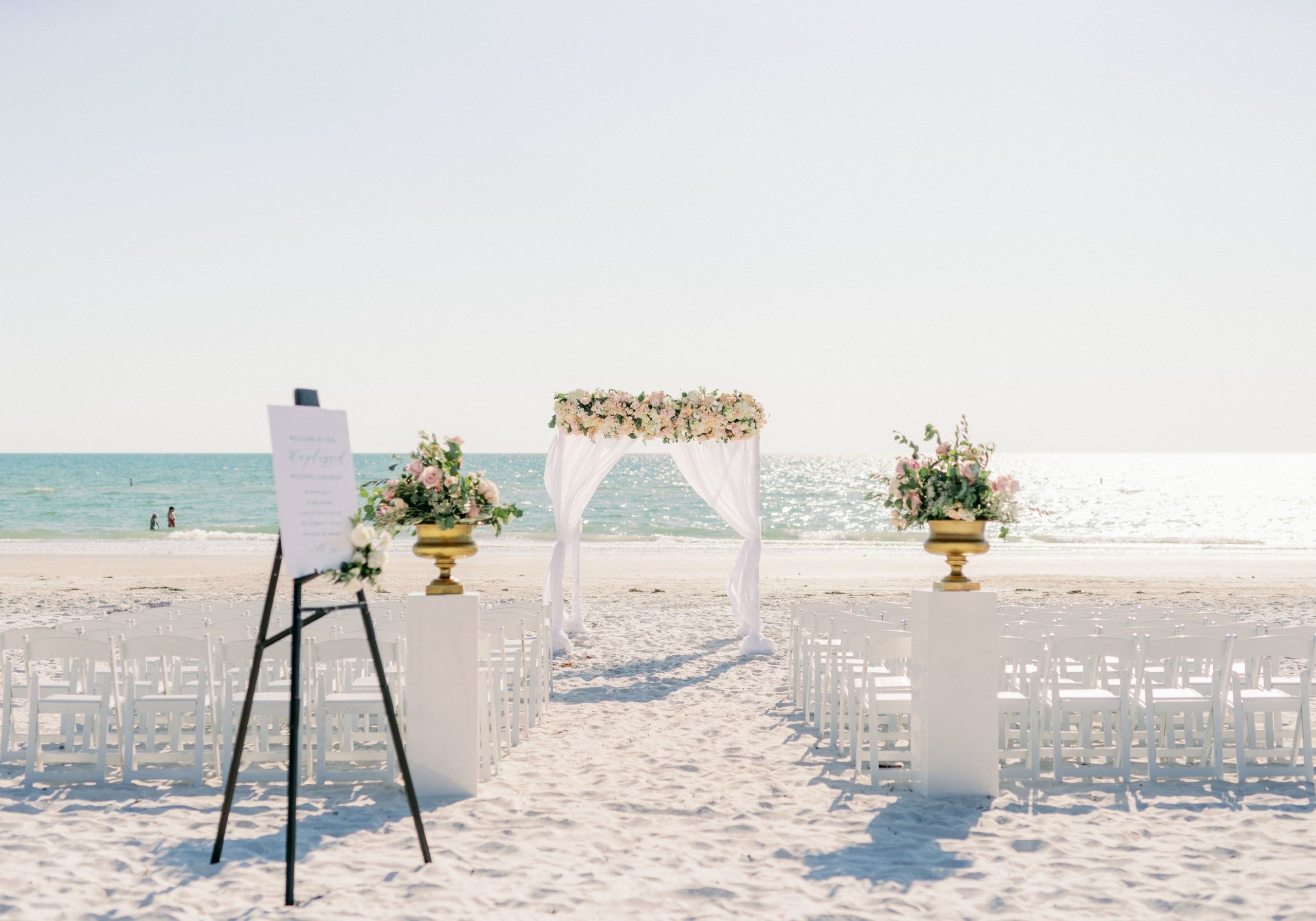 Beach wedding ceremony setup with an arch, floral arrangements, and chairs facing the ocean under a clear blue sky.