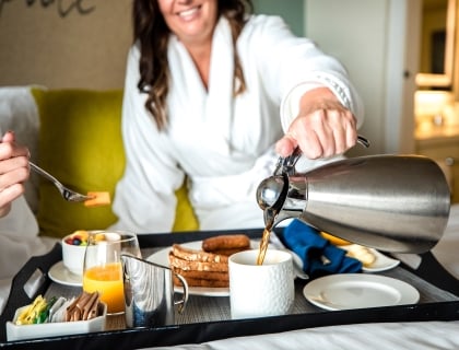 two women enjoying room service, one woman is pouring coffee and the other is eating fruit