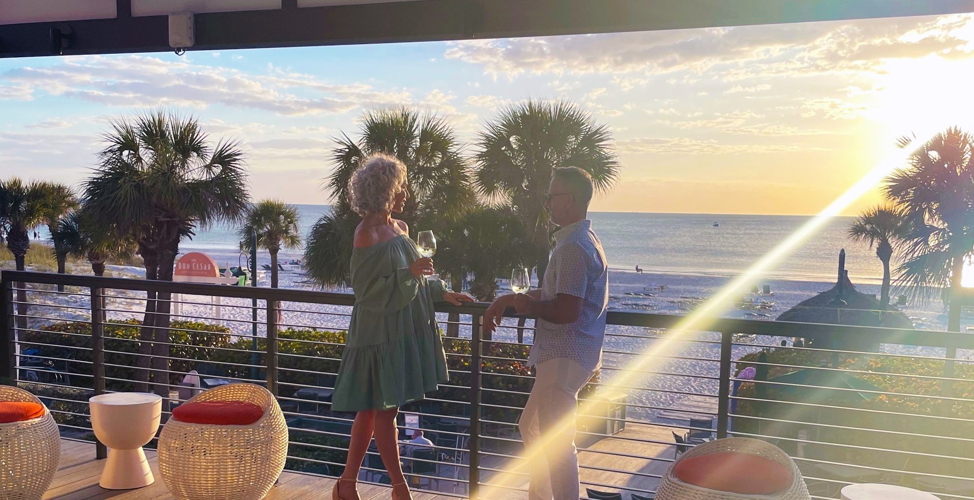 A couple stands on a balcony, gazing at the beach and ocean waves under a clear blue sky.