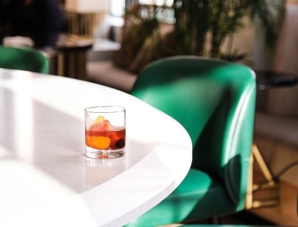 A glass of whiskey on a table with green chairs in the background.