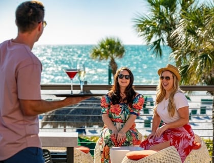 two women being served drinks on a balcony overlooking the ocean