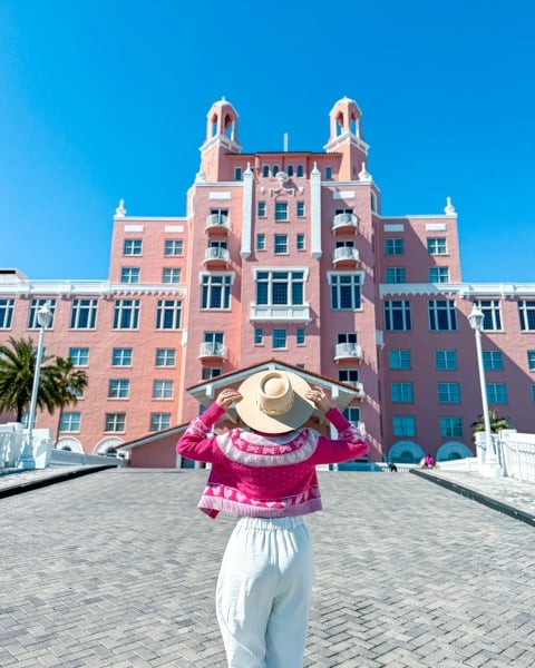 a woman wearing a white outfit and bright pink jacket holding her hat in front of The Don CeSar