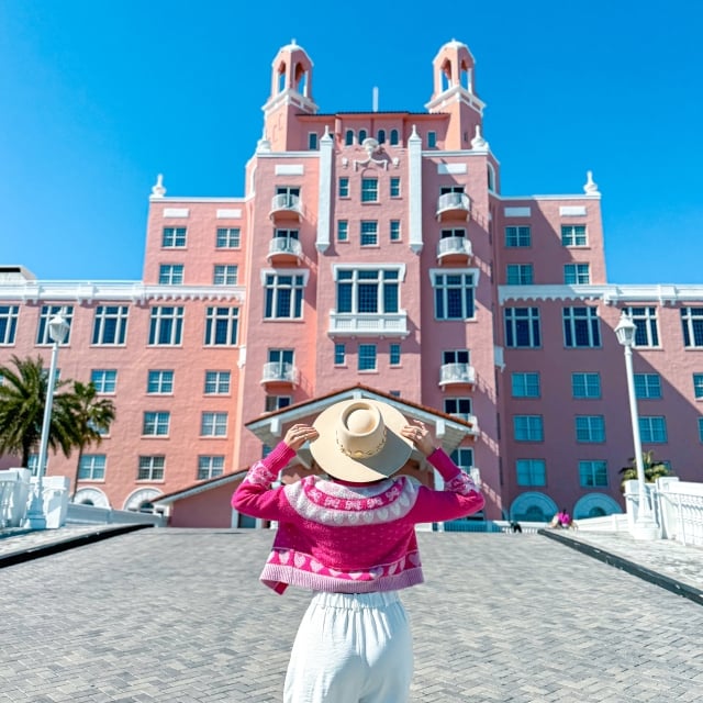 a woman wearing a white outfit and bright pink jacket holding her hat in front of The Don CeSar