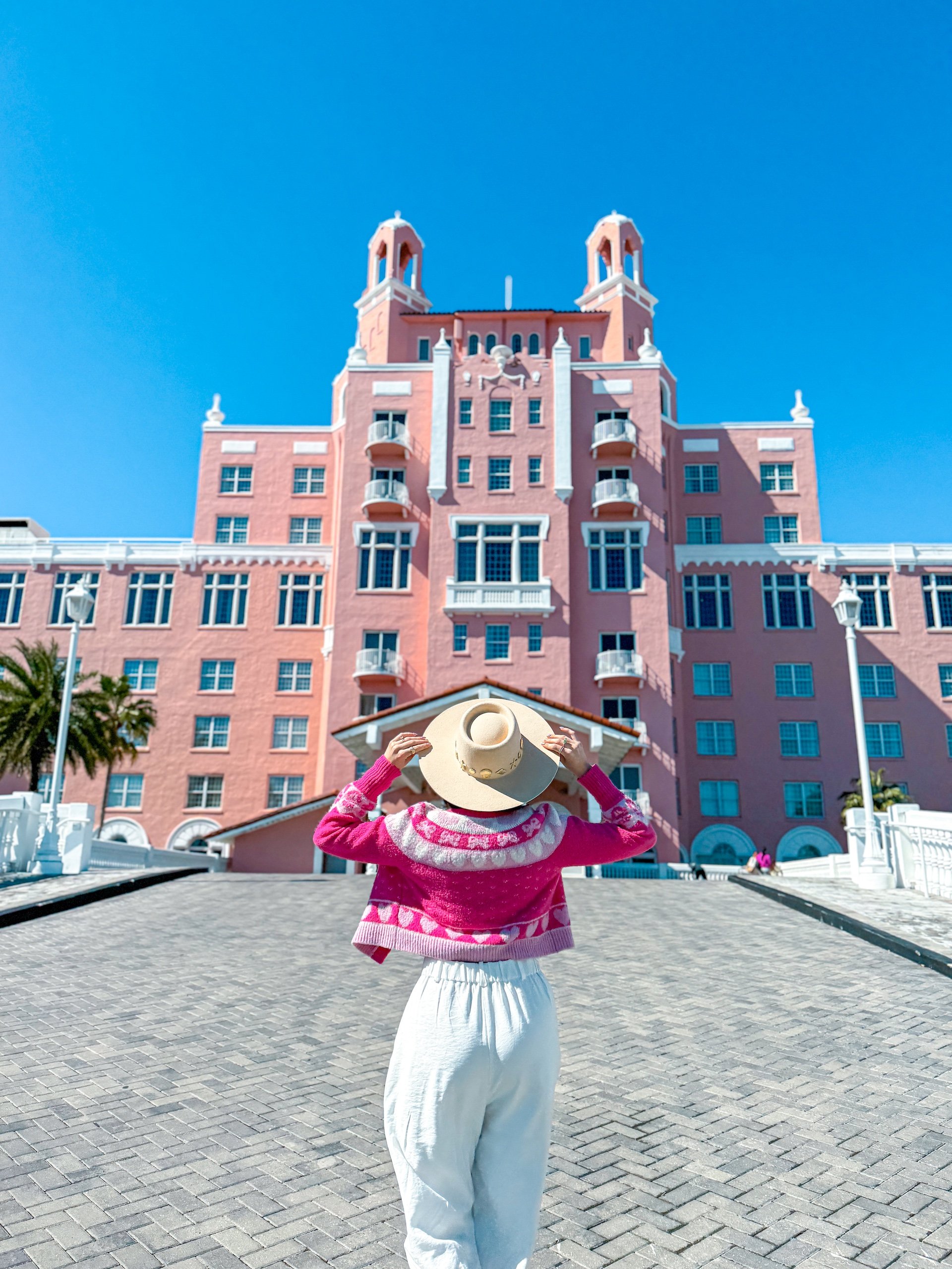 a woman wearing a white outfit and bright pink jacket holding her hat in front of The Don CeSar