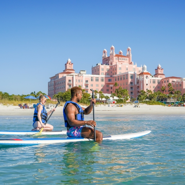 a couple paddleboarding in the ocean with the Don CeSar Hotel in the background