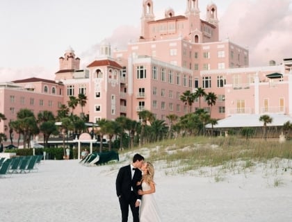 A bride and groom stand on the beach, smiling, with a pink hotel in the background.