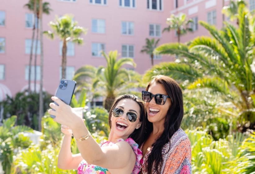 Two women smiling and taking a selfie in front of a pink hotel, capturing a fun moment together.