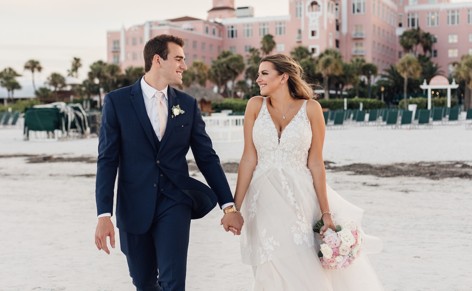 Bride and groom stroll hand in hand along the beach, with a hotel in the background, capturing a romantic moment.