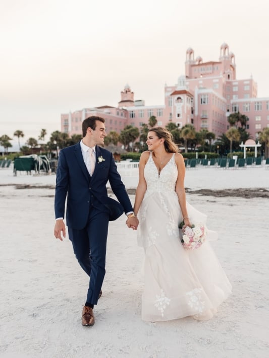 Bride and groom stroll hand in hand along the beach, with a hotel in the background, capturing a romantic moment.