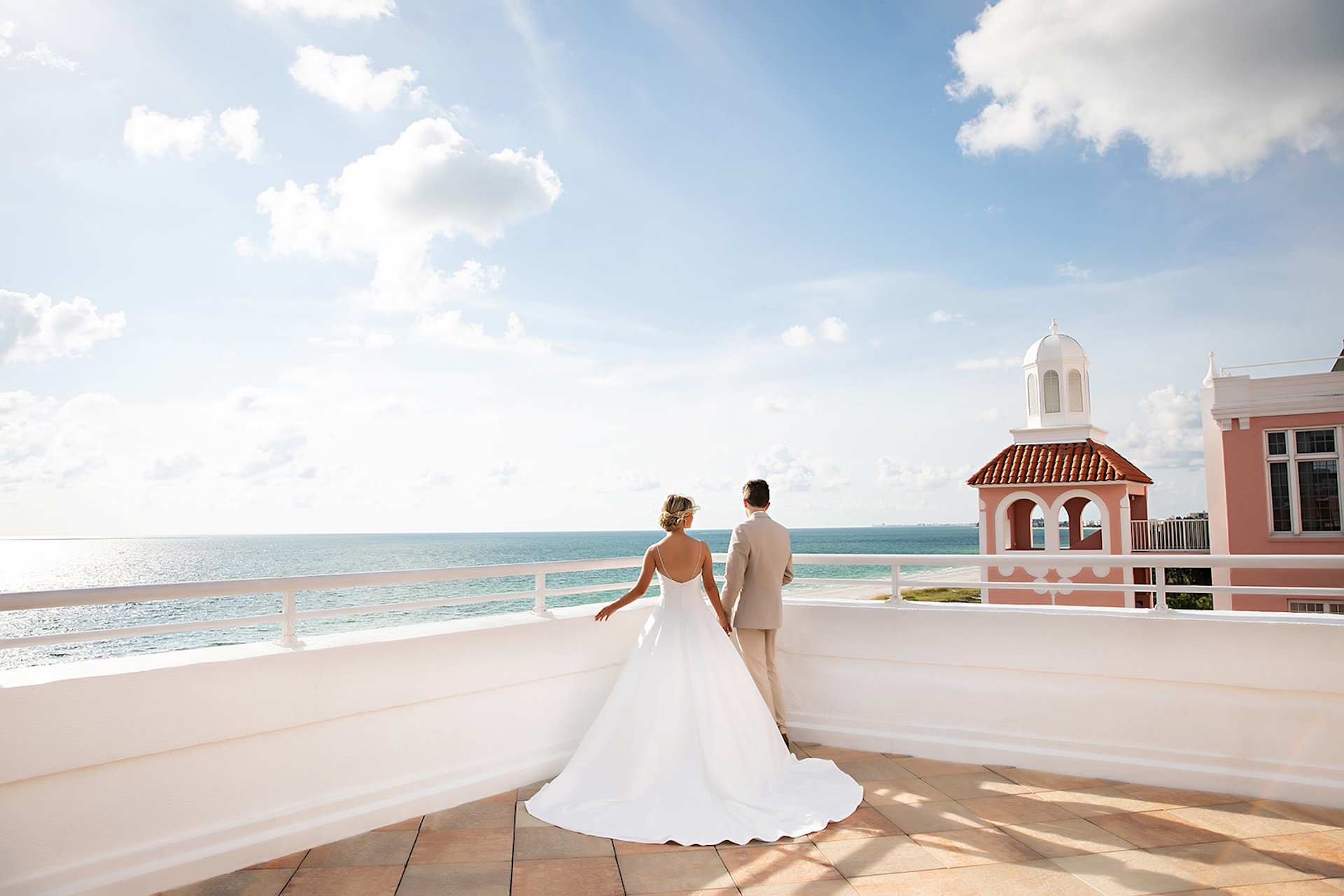 A bride and groom stand on a balcony, gazing at the ocean, surrounded by a romantic atmosphere.