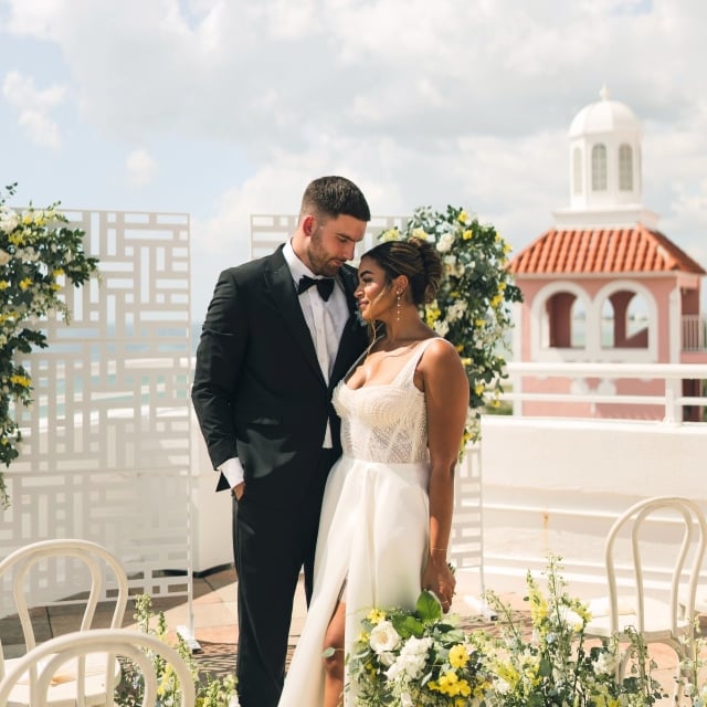 A bride and groom surrounded by flowers posing on a veranda