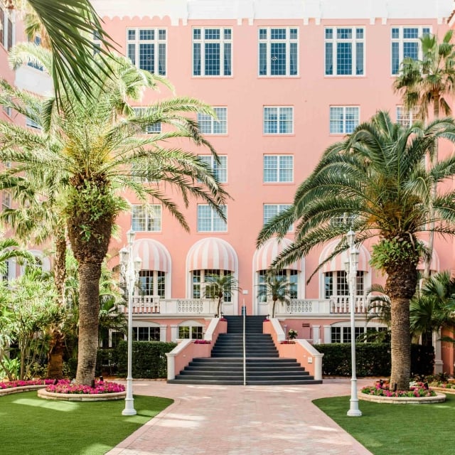 a courtyard surrounded by a pink hotel with large stair case and palm trees