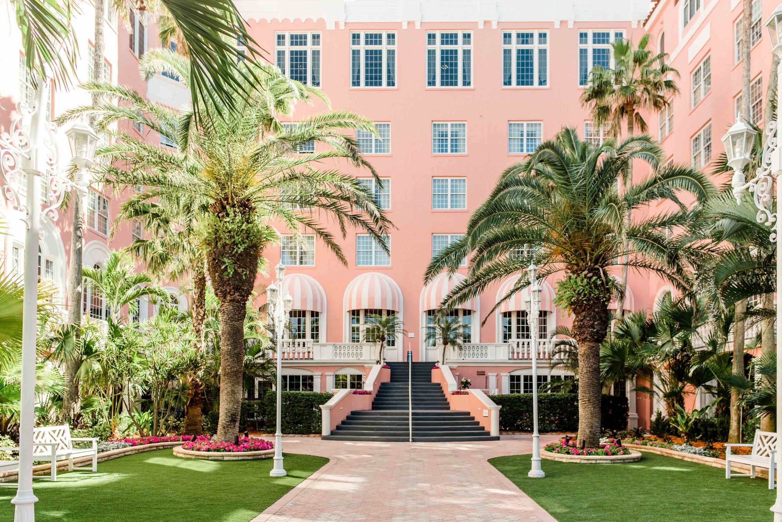 a courtyard surrounded by a pink hotel with large stair case and palm trees