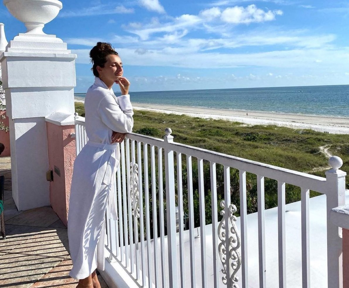 A woman in a white robe stands on a balcony, gazing out at the vast ocean below.